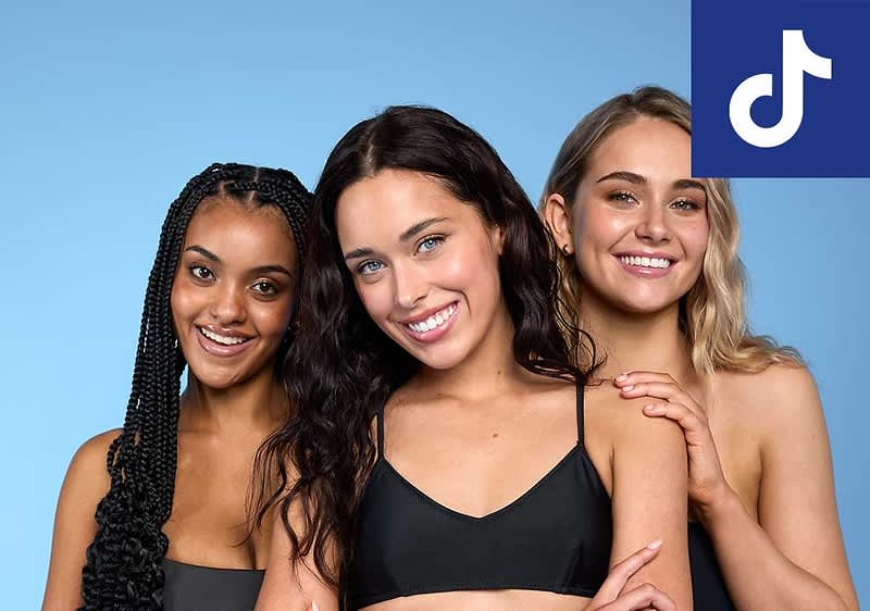 Drei junge Frauen in schwarzer Badekleidung stehen nebeneinander vor hellblauem Hintergrund, die mittlere Frau hat dunkelbraune, wellige Haare, die linke geflochtene lange Haare und die rechte schulterlange blonde Haare, oben rechts das weiße TikTok-Logo auf blauem Quadrat ohne weiteren Hintergrund