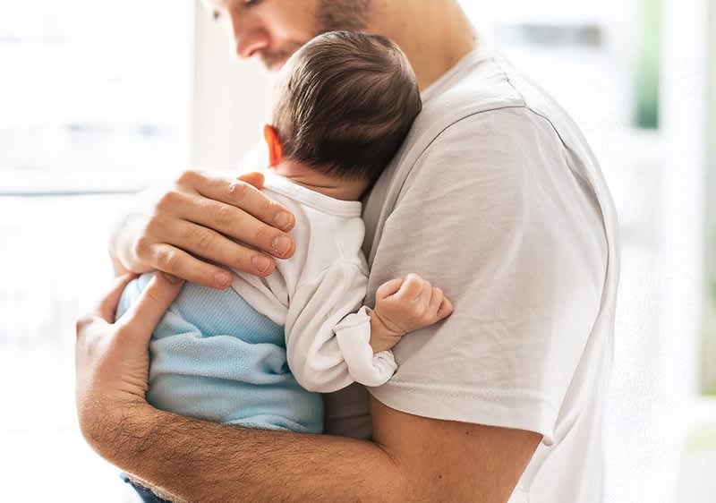 Ein Mann in einem weißen T-Shirt hält ein Baby im Arm, das einen weißen Langarmbody und eine hellblaue Hose trägt, das Baby liegt mit dem Kopf auf der Schulter des Mannes und seine Hand ist leicht zur Faust geballt, die Umgebung ist hell und unscharf im Hintergrund.