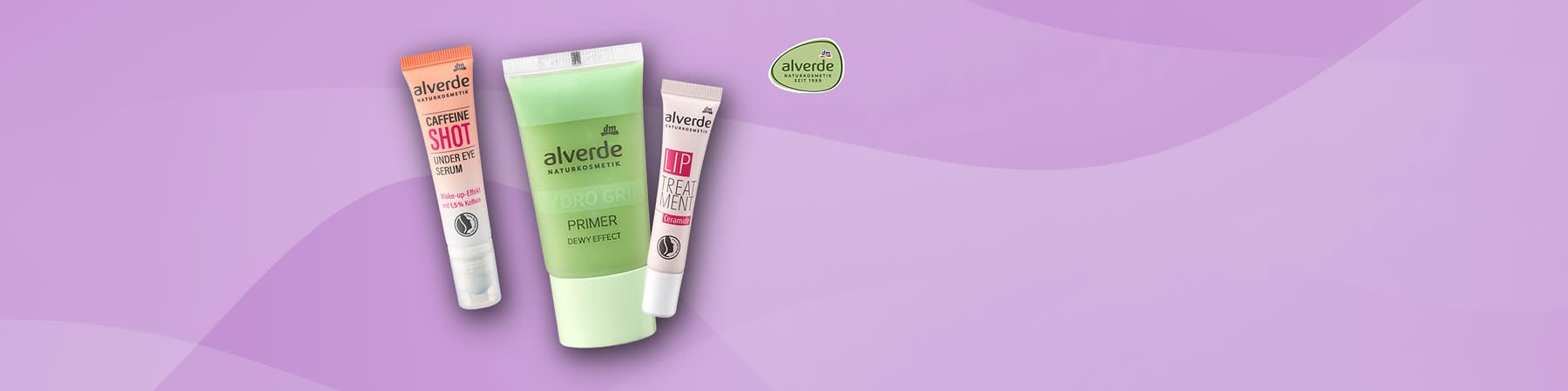 Drei alverde Naturkosmetik-Produkte vor fliederfarbenem Hintergrund: links eine orange-weiße Tube mit der Aufschrift „CAFFEINE SHOT UNDER EYE SERUM“, Mitte eine grün-weiße Tube mit der Aufschrift „alverde NATURKOSMETIK HYDRO GREEN PRIMER DEWY EFFECT“, rechts eine weiße Tube mit rosa Aufschrift „alverde LIP TREATMENT“, oben rechts das runde alverde-Logo ohne weiteren Text