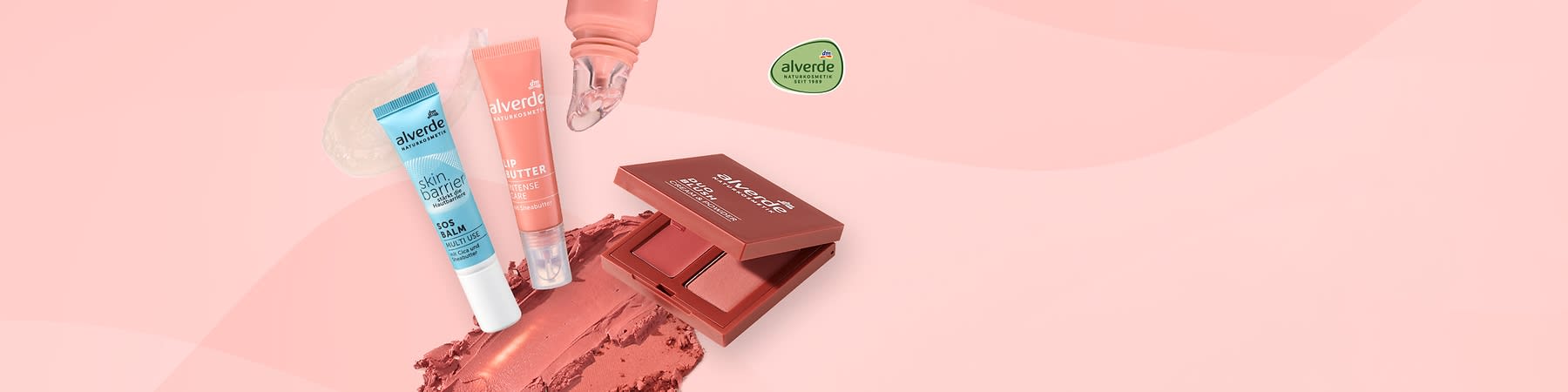 Drei alverde-Kosmetikprodukte auf roséfarbenem Hintergrund: links ein hellblaues Tube skin barrier SOS balm, mittig ein roséfarbenes Tube lip butter intense care, rechts ein offenes bronzefarbenes Rouge-Döschen mit zwei Farbtönen und Produktproben als Creme- und Puderstriche im Vordergrund, alverde-Logo oben rechts
