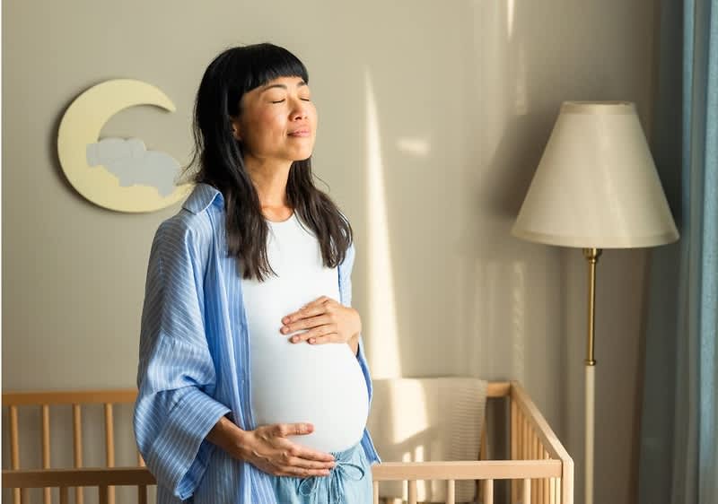 Schwangere Frau steht in einem Kinderzimmer und hält mit beiden Händen ihren freiliegenden Babybauch, im Hintergrund ein Babybett, eine Wanddekoration mit Mondsymbol und eine Stehlampe
