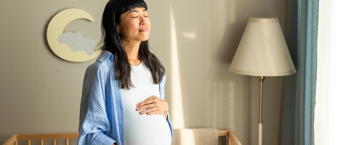 Schwangere Frau steht in einem Kinderzimmer und hält mit beiden Händen ihren freiliegenden Babybauch, im Hintergrund ein Babybett, eine Wanddekoration mit Mondsymbol und eine Stehlampe