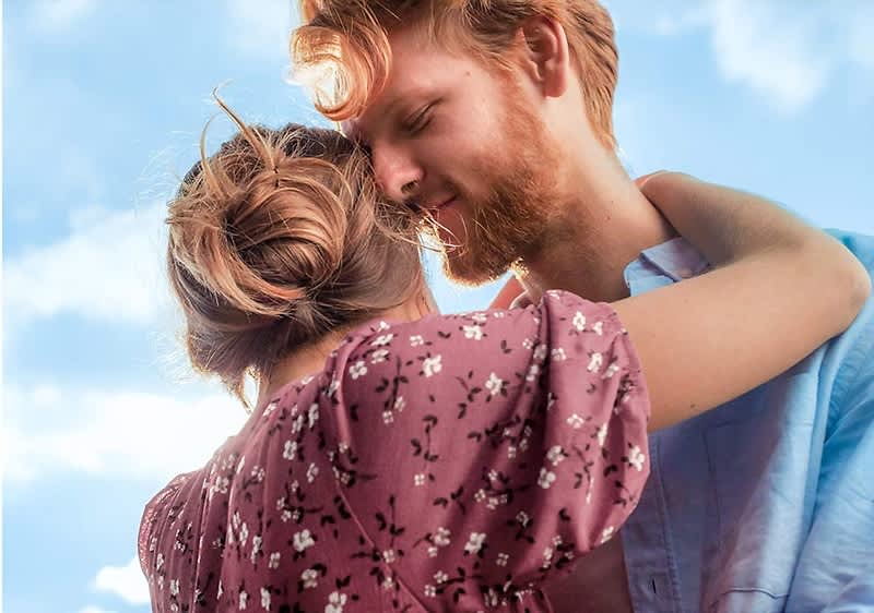 Eine Frau mit braunen Haaren, die zu einem lockeren Knoten gebunden sind, trägt ein rotes Kleid mit weißen Blumenmustern und umarmt einen Mann, dessen Gesicht unscharf ist, während er ein hellblaues Hemd trägt, im Hintergrund ist ein blauer Himmel mit weißen Wolken sichtbar