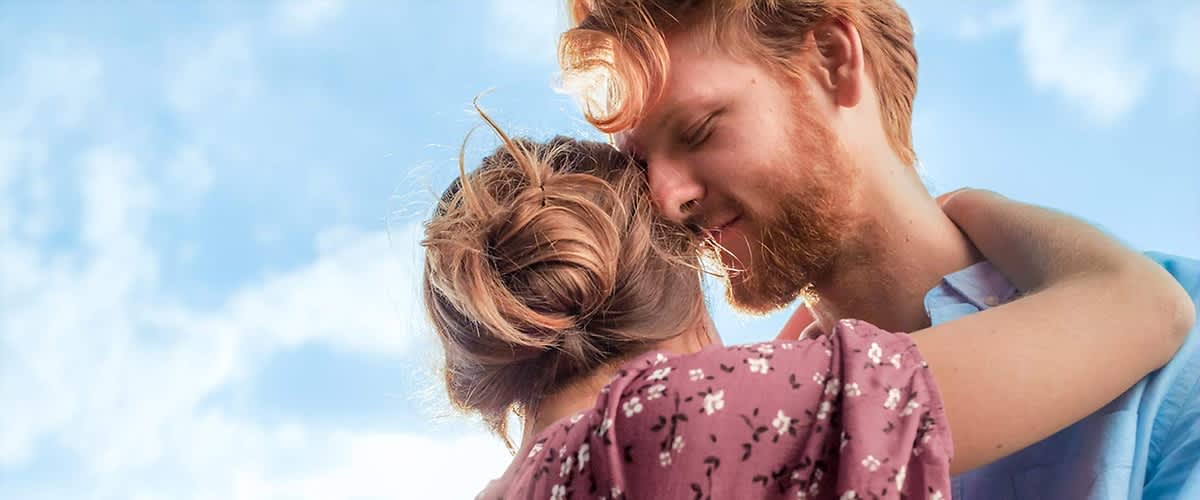 Eine Frau mit braunen Haaren, die zu einem lockeren Knoten gebunden sind, trägt ein rotes Kleid mit weißen Blumenmustern und umarmt einen Mann, dessen Gesicht unscharf ist, während er ein hellblaues Hemd trägt, im Hintergrund ist ein blauer Himmel mit weißen Wolken sichtbar