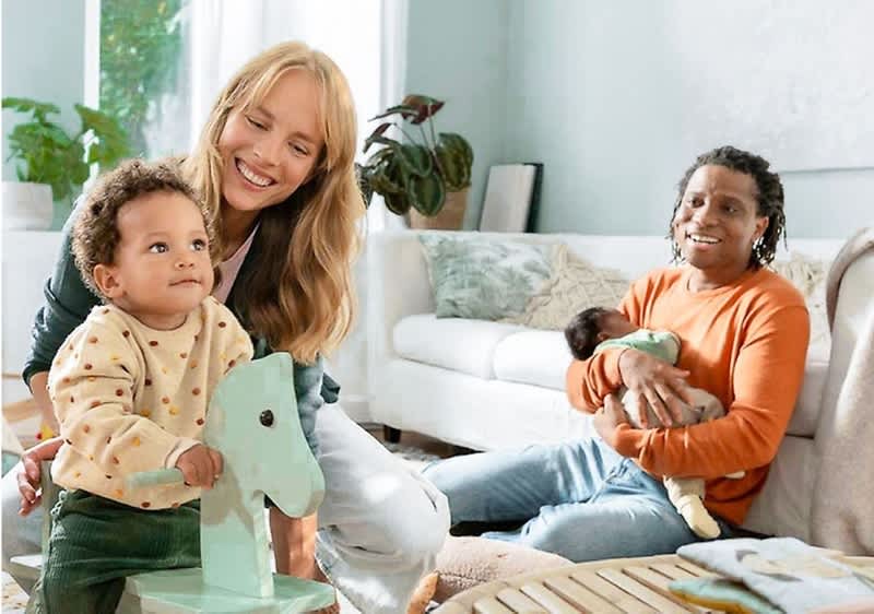 In einem hellen Wohnzimmer mit grünen Wänden und einem großen Fenster sitzt eine Person mit einem orangefarbenen Pullover auf einem Teppich und hält ein Baby im Arm, während ein Kleinkind in einem gepunkteten Pullover auf einem mintgrünen Schaukelpferd sitzt und eine weitere Person mit langen blonden Haaren hinter dem Kleinkind kniet, im Hintergrund sind ein weißes Sofa und eine grüne Zimmerpflanze zu sehen