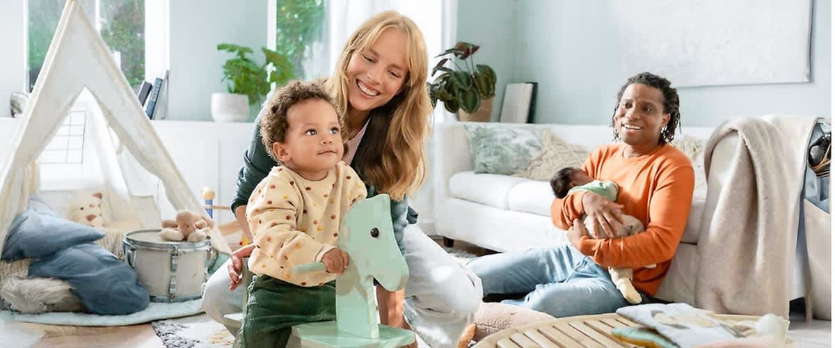 In einem hellen Wohnzimmer mit grünen Wänden und einem großen Fenster sitzt eine Person mit einem orangefarbenen Pullover auf einem Teppich und hält ein Baby im Arm, während ein Kleinkind in einem gepunkteten Pullover auf einem mintgrünen Schaukelpferd sitzt und eine weitere Person mit langen blonden Haaren hinter dem Kleinkind kniet, im Hintergrund sind ein weißes Sofa und eine grüne Zimmerpflanze zu sehen