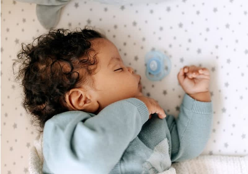 Schlafendes Baby in blaugrünem Strickoberteil liegt auf einer hellen Matratze mit kleinen Sternen, neben dem Baby liegt ein blauer Schnuller