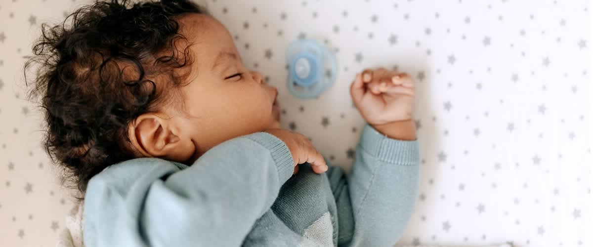 Schlafendes Baby in blaugrünem Strickoberteil liegt auf einer hellen Matratze mit kleinen Sternen, neben dem Baby liegt ein blauer Schnuller