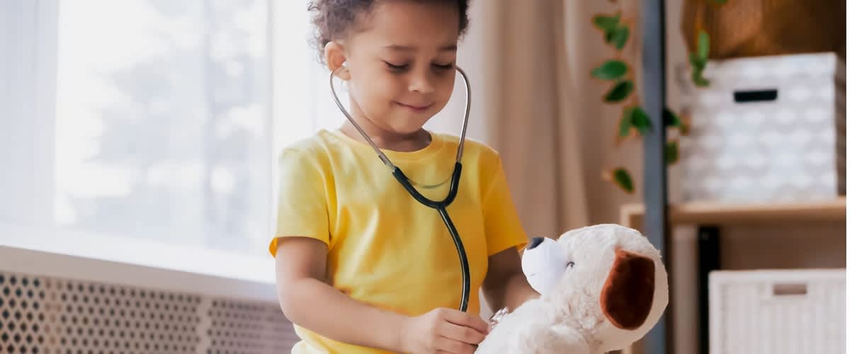 Kindesgröße sitzt mit Spielzeug-Stethoskop an Ohren und hört mit dem Stethoskop an einem weißen Plüschtierhund, trägt gelbes T-Shirt und blaue Hose