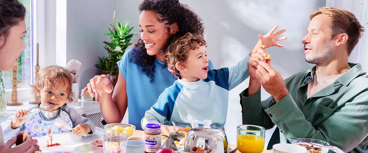 Familie sitzt am Frühstückstisch; Kind in blau-weißem Shirt reicht die Hand nach einem Elternteil, auf dem Tisch Orangensaft, Teekanne, Müsli, Obst und ein Marmeladenglas sichtbar