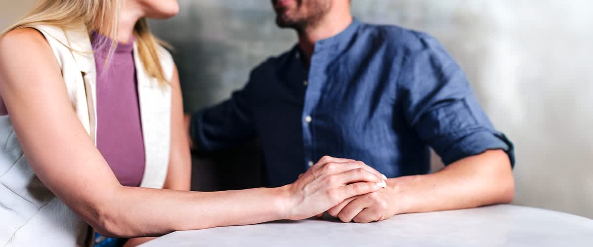 Ein Mann und eine Frau sitzen an einem Tisch und halten sich die Hände, der Mann trägt ein blaues Hemd und die Frau ein weißes Oberteil, beide sind nur teilweise sichtbar, oben rechts steht der Schriftzug fertia health in dunkelblauer Farbe mit einem kleinen stilisierten Vogel daneben