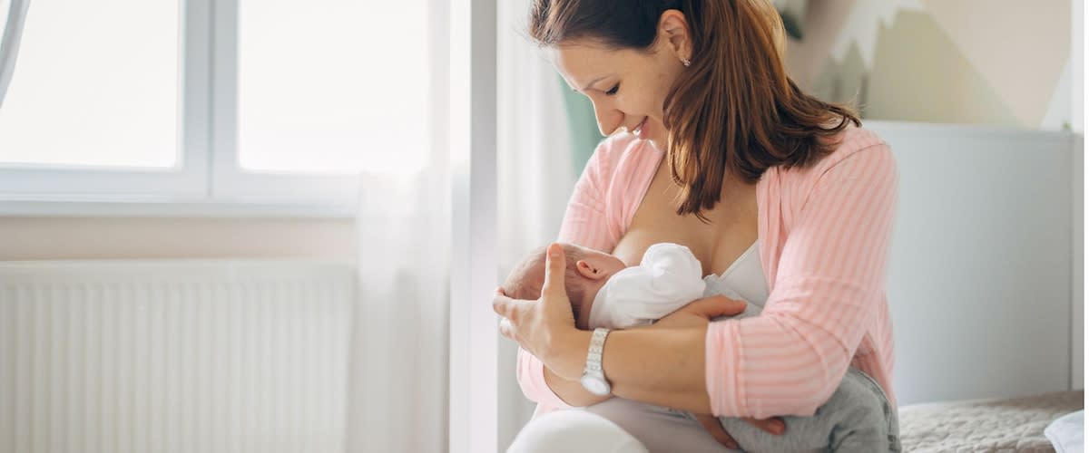 Frau sitzt auf Bettkante und hält ein schlafendes Baby im Arm, beide in heller Alltagskleidung, im Hintergrund Fenster und Kinderzimmermöbel