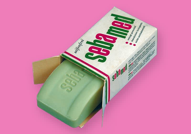 Eine grüne Seife der Marke Sebamed liegt halb aus ihrer weißen Verpackung heraus, die Verpackung ist mit dem grünen und roten Schriftzug sebamed bedruckt und hat einen pinken Hintergrund