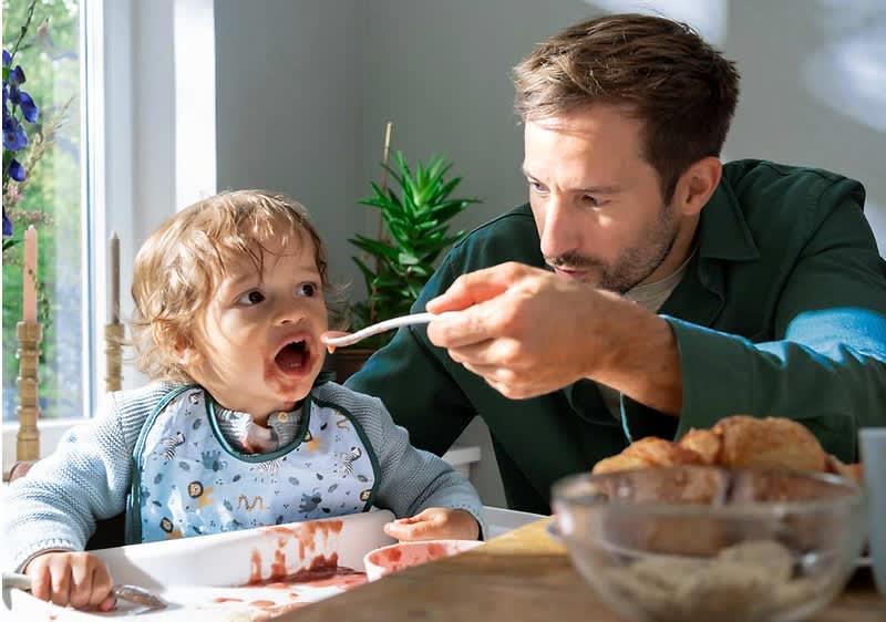 Ein Kleinkind sitzt in einem Hochstuhl und trägt einen Lätzchen mit bunten Motiven, während ein Erwachsener ihm mit einem Löffel Essen reicht, auf dem Tisch stehen ein Teller mit Croissants und eine Schale mit Müsli, im Hintergrund sind eine grüne Pflanze und ein Fenster mit Tageslicht zu sehen