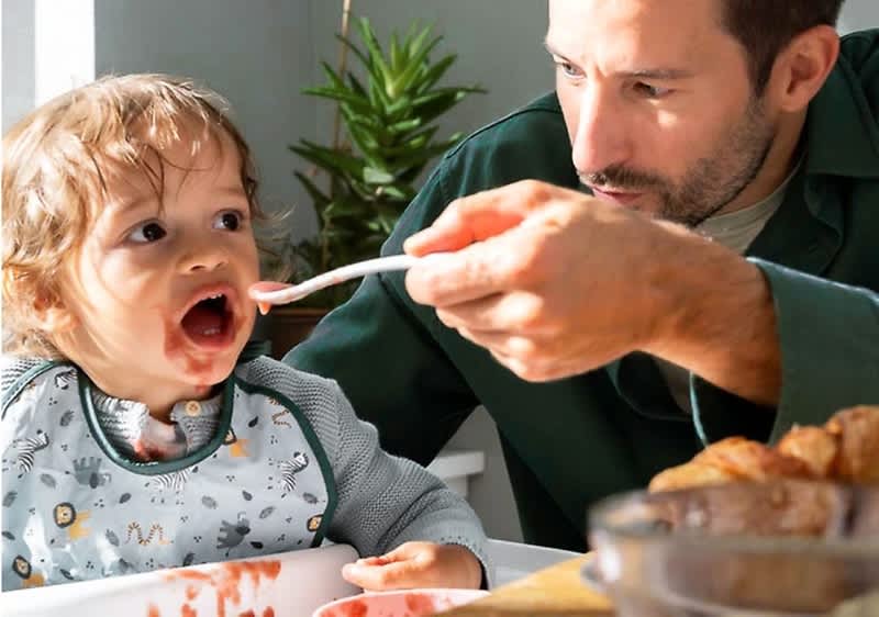 Ein Kleinkind sitzt in einem Hochstuhl und trägt einen Lätzchen mit bunten Motiven, während ein Erwachsener ihm mit einem Löffel Essen reicht, auf dem Tisch stehen ein Teller mit Croissants und eine Schale mit Müsli, im Hintergrund sind eine grüne Pflanze und ein Fenster mit Tageslicht zu sehen