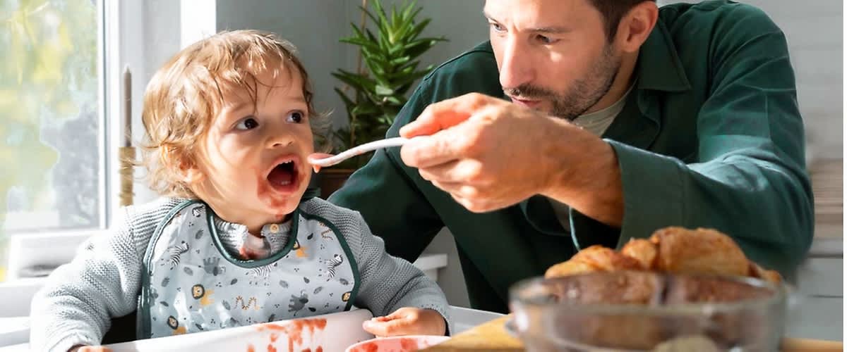Ein Kleinkind sitzt in einem Hochstuhl und trägt einen Lätzchen mit bunten Motiven, während ein Erwachsener ihm mit einem Löffel Essen reicht, auf dem Tisch stehen ein Teller mit Croissants und eine Schale mit Müsli, im Hintergrund sind eine grüne Pflanze und ein Fenster mit Tageslicht zu sehen