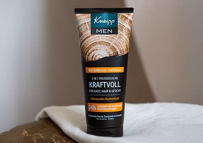 Eine schwarze Tube mit grünen Akzenten der Marke Kneipp MEN steht auf einem beigen Fliesenboden, darauf steht Haut & Haar 2 in 1 Dusche STARTKLAR mit den Duftnoten Lemongras und Guarana, darunter der Hinweis Natürliches ätherisches Öl, daneben liegt ein Stück beige Seife.