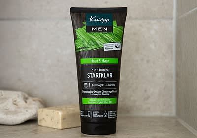 Eine schwarze Tube mit der Aufschrift Kneipp MEN und darunter Bio Zedernuss + Panthenol steht auf einem weißen Handtuch, die Tube enthält eine 3 in 1 Pflegedusche mit der Bezeichnung KRAFTVOLL für Haut, Haar & Gesicht, darunter steht Aktivierendes Duscherlebnis und 24h bewahrt die natürliche Hautfeuchtigkeit, die Tube liegt auf einem Holztisch und hat ein braunes Muster im oberen Bereich.