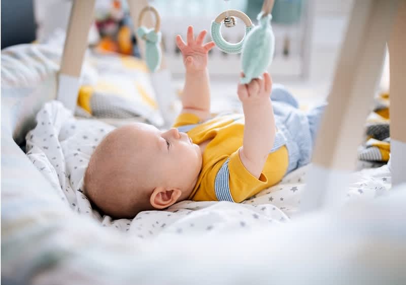 Säugling liegt auf dem Rücken in einer gepolsterten Babywippe und greift mit beiden Händen nach hängenden Spielzeugen an einem Spielbogen