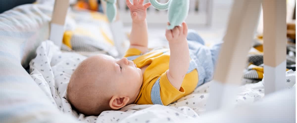 Säugling liegt auf dem Rücken in einer gepolsterten Babywippe und greift mit beiden Händen nach hängenden Spielzeugen an einem Spielbogen