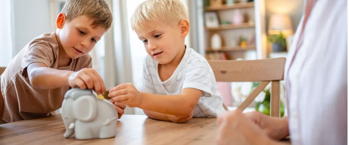 Zwei kleine Kinder legen Münzen in ein graues Sparschwein auf einem Tisch, rechts eine Erwachsene sitzt beobachtend am Tischstuhl