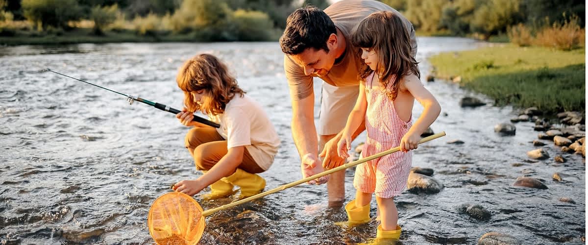 Vater kniet im flachen Fluss und hilft zwei Kindern in Gummistiefeln beim Fischen mit Netz und Angel, sonnenbeschienene Uferbäume im Hintergrund