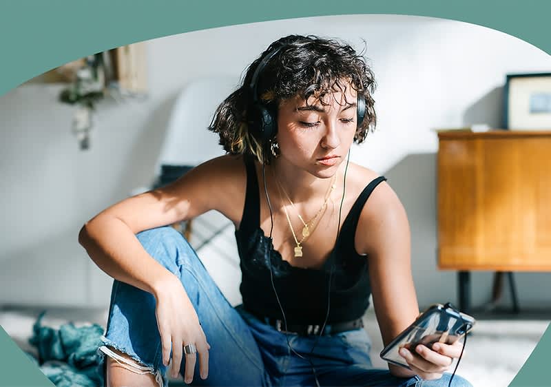 Eine Frau mit lockigem, schulterlangem Haar sitzt auf dem Boden und trägt ein schwarzes Top und blaue Jeans, sie hält ein Smartphone in der rechten Hand und trägt große Kopfhörer, im Hintergrund sind ein Holzmöbelstück und ein gerahmtes Bild zu sehen, die Gesichtszüge der Frau sind unscharf gemacht.