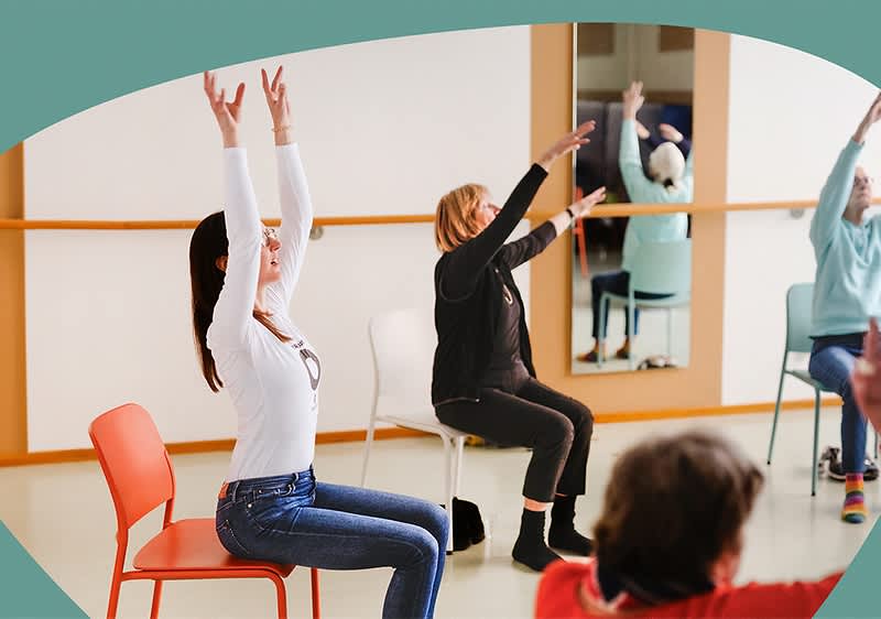 Zwei Frauen sitzen auf Stühlen in einem Raum mit hellem Boden und weißen Wänden; die Frau links trägt ein weißes Langarmshirt, blaue Jeans und rote Socken und beugt sich nach vorne, während die Frau rechts ebenfalls ein weißes Langarmshirt und blaue Jeans trägt und aufrecht sitzt; im Hintergrund ist eine weitere Person in einem roten Pullover und dunkler Hose auf einem weißen Stuhl zu sehen, deren Gesicht unscharf ist