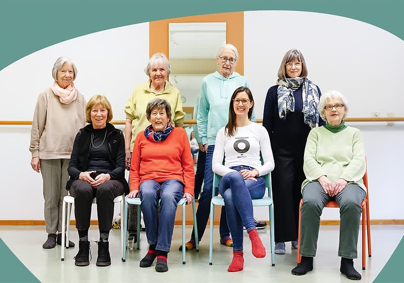 Eine Gruppe von sieben Personen sitzt und steht in einem Raum mit hellgrünem Boden und einer weißen Wand, die durch einen orangefarbenen Rahmen unterbrochen wird, alle tragen lässige Kleidung in verschiedenen Farben wie Rot, Grün, Gelb und Schwarz, und einige haben Schals um den Hals, während die Gesichter der Personen unscharf sind.