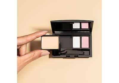 Eine Hand mit hellrosa lackierten Nägeln hält einen rechteckigen, beigen Lidschatten vor einer geöffneten schwarzen Lidschattenpalette, die zwei weitere Lidschattenfarben in Weiß und Rosa sowie einen kleinen Applikator enthält, der auf einem beigen Hintergrund liegt