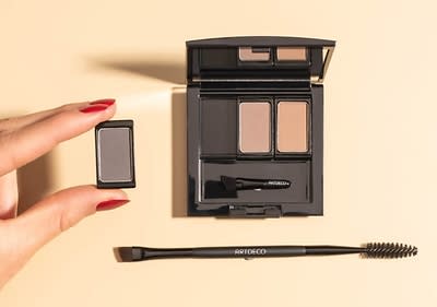 Eine Hand mit roten Fingernägeln hält einen kleinen rechteckigen Lidschatten in Grau, daneben liegt eine geöffnete schwarze Lidschattenpalette mit zwei Farben, Grau und Braun, und einem kleinen Applikator mit der Aufschrift ARTDECO, darunter liegt ein schwarzer doppelseitiger Augenbrauenpinsel mit der Aufschrift ARTDECO auf einem hellbeigen Hintergrund