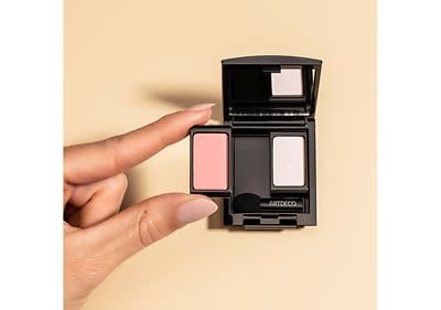 Eine Hand mit hellrosa lackierten Nägeln hält einen kleinen rechteckigen Lidschatten in Rosa, daneben eine geöffnete schwarze Lidschattenpalette von Artdeco mit zwei Fächern, eines leer und das andere enthält einen weißen Lidschatten, auf der Palette ist der Schriftzug ARTDECO zu sehen.