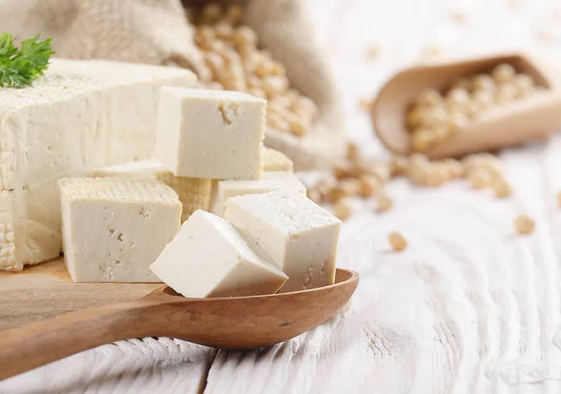 Ein Holzlöffel mit mehreren weißen Tofuwürfeln liegt auf einem hellen Holztisch, daneben ein größerer Block Tofu, im Hintergrund sind ein Jutesack und ein Holzlöffel mit Sojabohnen zu sehen, ein grünes Blatt liegt auf dem Tofu-Block.