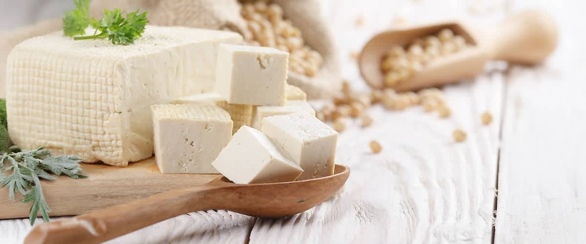 Ein Holzlöffel mit mehreren weißen Tofuwürfeln liegt auf einem hellen Holztisch, daneben ein größerer Block Tofu, im Hintergrund sind ein Jutesack und ein Holzlöffel mit Sojabohnen zu sehen, ein grünes Blatt liegt auf dem Tofu-Block.