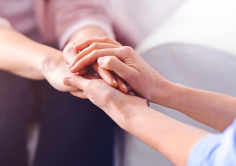 Drei Personen sitzen an einem Tisch in einem hellen Raum mit großen Fenstern, die zwei Frauen tragen lässige Kleidung und schauen auf einen Laptop, während der Mann in einem weißen Hemd Notizen macht, auf dem Tisch liegen ein Stethoskop und ein Kugelschreiber, oben rechts im Bild steht der Schriftzug fertia health in dunkelblauer Farbe mit einem kleinen Vogel-Symbol daneben.