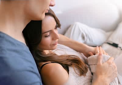 Ein Mann mit kurzem braunem Haar und einem blauen T-Shirt hält eine Frau mit langen braunen Haaren, die einen cremefarbenen Strickpullover trägt, in einer liebevollen Umarmung, während sie auf einem Sofa sitzen; oben rechts im Bild steht der Schriftzug fertia health in dunkelblauer Schrift mit einem kleinen stilisierten Vogel daneben.