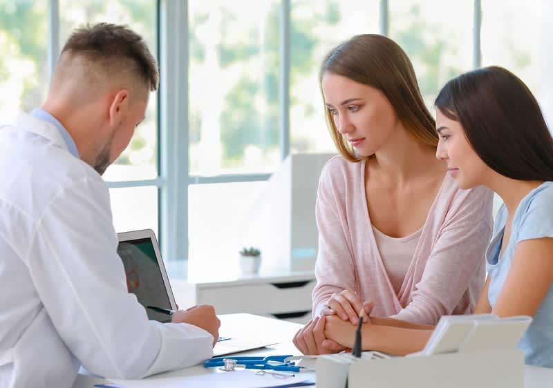 Drei Personen sitzen an einem Tisch in einem hellen Raum mit großen Fenstern, die zwei Frauen tragen lässige Kleidung und schauen auf einen Laptop, während der Mann in einem weißen Hemd Notizen macht, auf dem Tisch liegen ein Stethoskop und ein Kugelschreiber, oben rechts im Bild steht der Schriftzug fertia health in dunkelblauer Farbe mit einem kleinen Vogel-Symbol daneben.