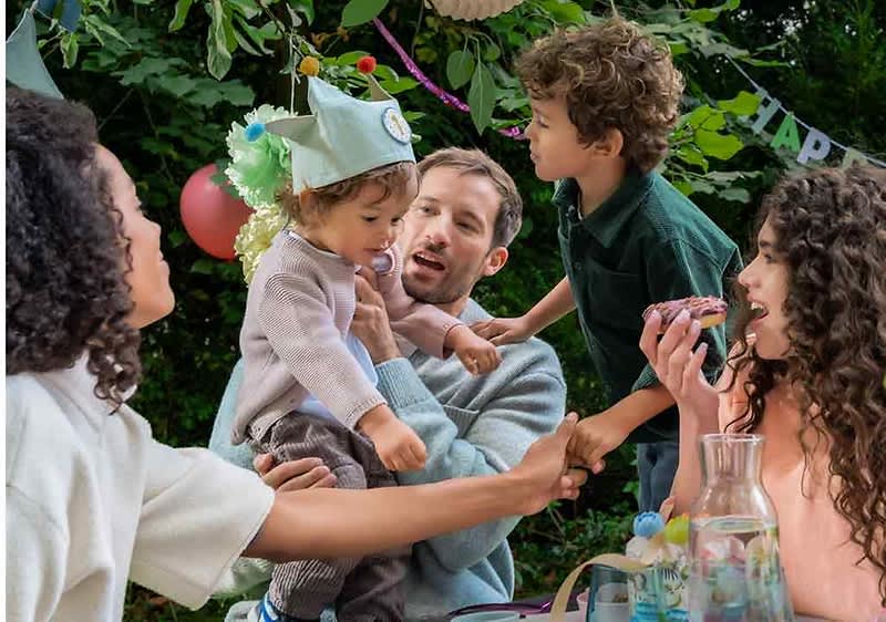 Eine Gruppe von Menschen sitzt im Freien an einem Tisch, der mit einer Glasflasche und einem Korb dekoriert ist, ein Kleinkind trägt einen Partyhut mit Pompons und wird von einem Erwachsenen gehalten, während ein weiteres Kind sich zu ihnen hinüberbeugt, im Hintergrund sind grüne Blätter und bunte Luftballons zu sehen, und ein Banner mit der Aufschrift HAPPY hängt zwischen den Bäumen
