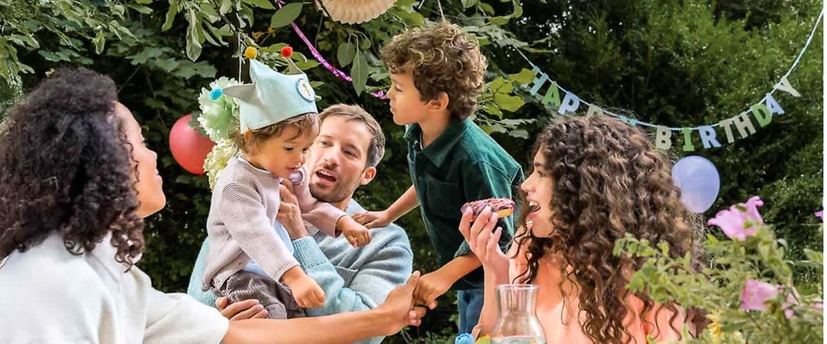 Eine Gruppe von Menschen sitzt im Freien an einem Tisch, der mit einer Glasflasche und einem Korb dekoriert ist, ein Kleinkind trägt einen Partyhut mit Pompons und wird von einem Erwachsenen gehalten, während ein weiteres Kind sich zu ihnen hinüberbeugt, im Hintergrund sind grüne Blätter und bunte Luftballons zu sehen, und ein Banner mit der Aufschrift HAPPY hängt zwischen den Bäumen
