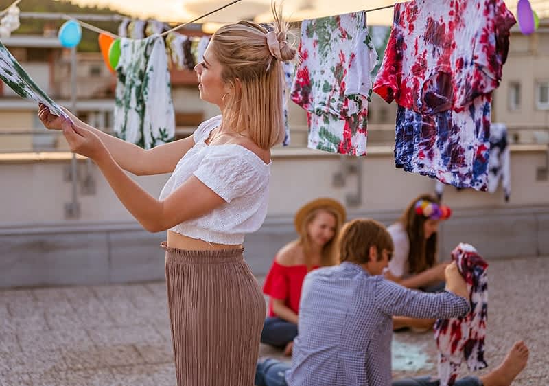 Eine Frau mit blonden Haaren, die zu einem Dutt gebunden sind, steht auf einer Dachterrasse und hängt bunte, batikgemusterte T-Shirts an eine Wäscheleine, während im Hintergrund zwei Personen mit verschwommenen Gesichtern auf dem Boden sitzen und ebenfalls T-Shirts betrachten; die Szene ist von einer sommerlichen Atmosphäre geprägt, mit bunten Luftballons, die an der Leine hängen, und einem Gebäude im Hintergrund.