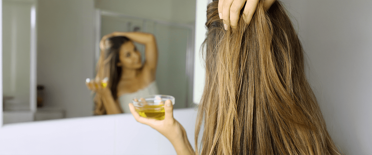 Eine Frau mit langen, braunen Haaren steht vor einem Spiegel und hält eine kleine Glasschale mit gelblicher Flüssigkeit in der rechten Hand, während sie mit der linken Hand ihre Haare berührt.