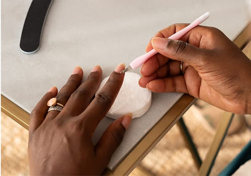 Zwei dunkelhäutige Hände, eine hält ein rosa Nagelhautschieber und bearbeitet die Nagelhaut der anderen Hand, die auf einem Tisch liegt, daneben liegt eine weiße runde Wattepadscheibe und eine schwarze Nagelfeile