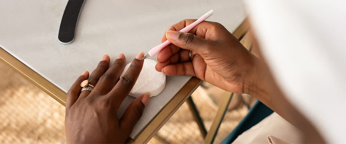 Zwei dunkelhäutige Hände, eine hält ein rosa Nagelhautschieber und bearbeitet die Nagelhaut der anderen Hand, die auf einem Tisch liegt, daneben liegt eine weiße runde Wattepadscheibe und eine schwarze Nagelfeile