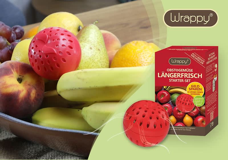 Eine braune Schale mit verschiedenen Früchten wie Trauben, Pfirsich, Birne, Banane und Orange steht auf einem Tisch, daneben eine rote Kugel mit Löchern und eine Verpackung mit der Aufschrift Wrappy Obst & Gemüse längerfrisch Starter-Set, Geld sparen, Frische bis zu 3x länger, Für Obst und Gemüse, Mit 2 Frischekugeln und einem Bild von verschiedenen Früchten auf der Verpackung.