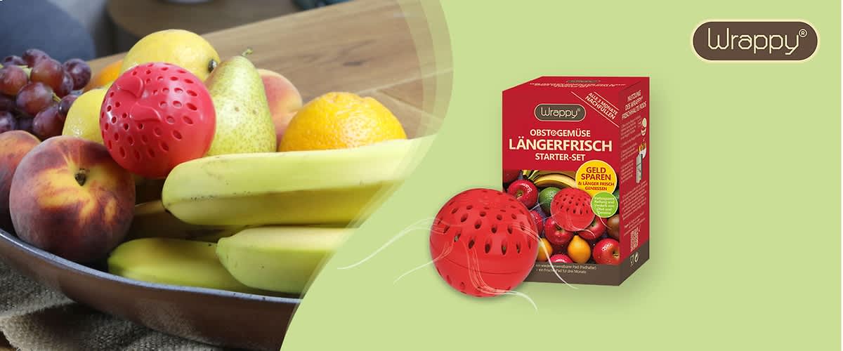 Eine braune Schale mit verschiedenen Früchten wie Trauben, Pfirsich, Birne, Banane und Orange steht auf einem Tisch, daneben eine rote Kugel mit Löchern und eine Verpackung mit der Aufschrift Wrappy Obst & Gemüse längerfrisch Starter-Set, Geld sparen, Frische bis zu 3x länger, Für Obst und Gemüse, Mit 2 Frischekugeln und einem Bild von verschiedenen Früchten auf der Verpackung.