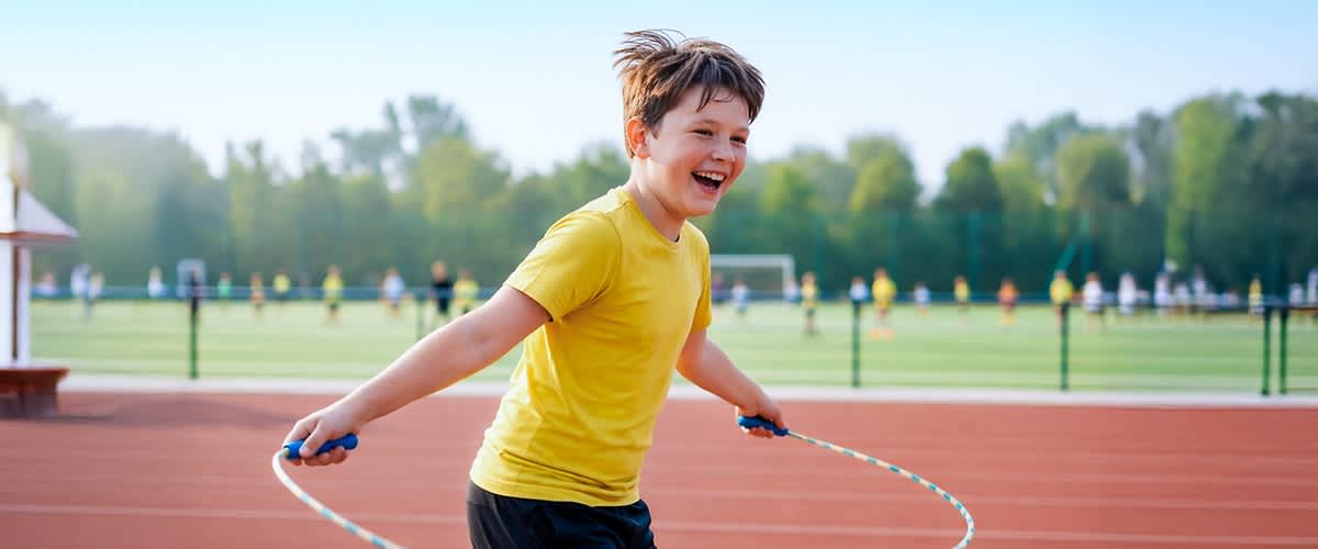 Junge in gelbem T‑Shirt und dunkler Sporthose springt auf einer Laufbahn mit einem Springseil, im Hintergrund Sportplatz mit Tor und weiteren Personen