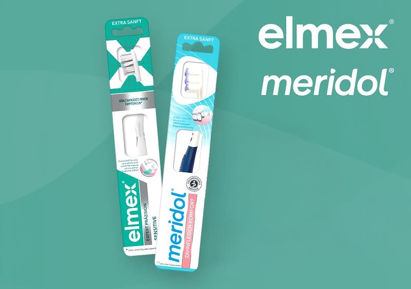 Zwei Zahnbürstenverpackungen stehen nebeneinander, die linke Verpackung ist von der Marke Elmex mit der Aufschrift Extra Sanft und zeigt eine weiße Zahnbürste, die rechte Verpackung ist von der Marke Meridol mit der Aufschrift Extra Sanft und zeigt eine Zahnbürste mit weißen und blauen Borsten, daneben befindet sich ein Recycling-Symbol aus drei grauen Pfeilen, oben rechts ist das grüne Elmex-Logo mit weißem Schriftzug zu sehen
