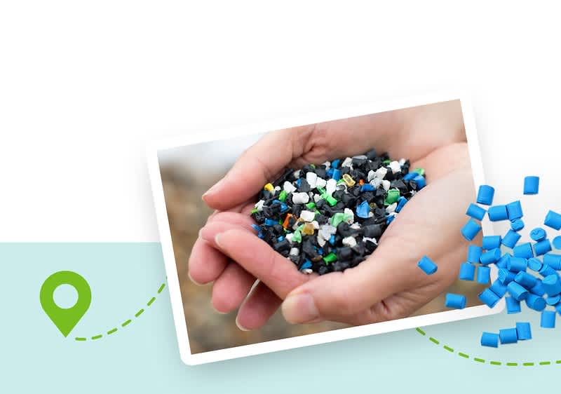 Eine Illustration zeigt eine Fotografie von weißen und schwarzen Kunststoffgranulaten nebeneinander, die auf einem hellblauen Hintergrund mit einem grünen Standort-Symbol und einer gestrichelten Linie platziert ist, einige Granulatstücke sind aus dem Bild herausgefallen und liegen rechts daneben.