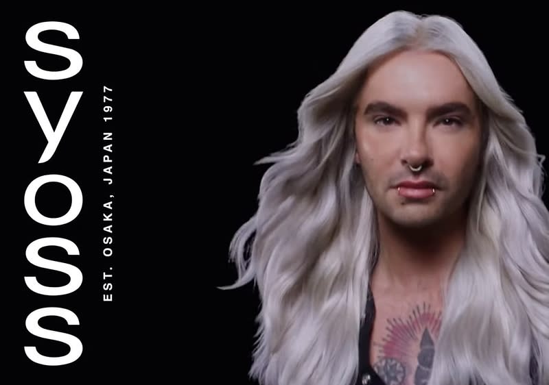 Bill Kaulitz mit langen, welligen, silberblonden Haaren und einem tätowierten Dekolleté vor einem schwarzen Hintergrund, links steht in weißer Schrift vertikal SYOSS und darunter EST. OSAKA, JAPAN 1977