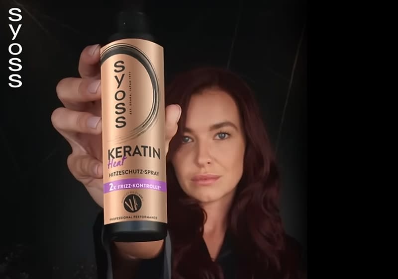 Eine Person mit langen, dunkelroten Haaren hält eine Flasche Syoss Keratin Heat Hitzeschutz-Spray in der Hand, daneben ist das Syoss-Logo zu sehen, der Hintergrund ist dunkel.